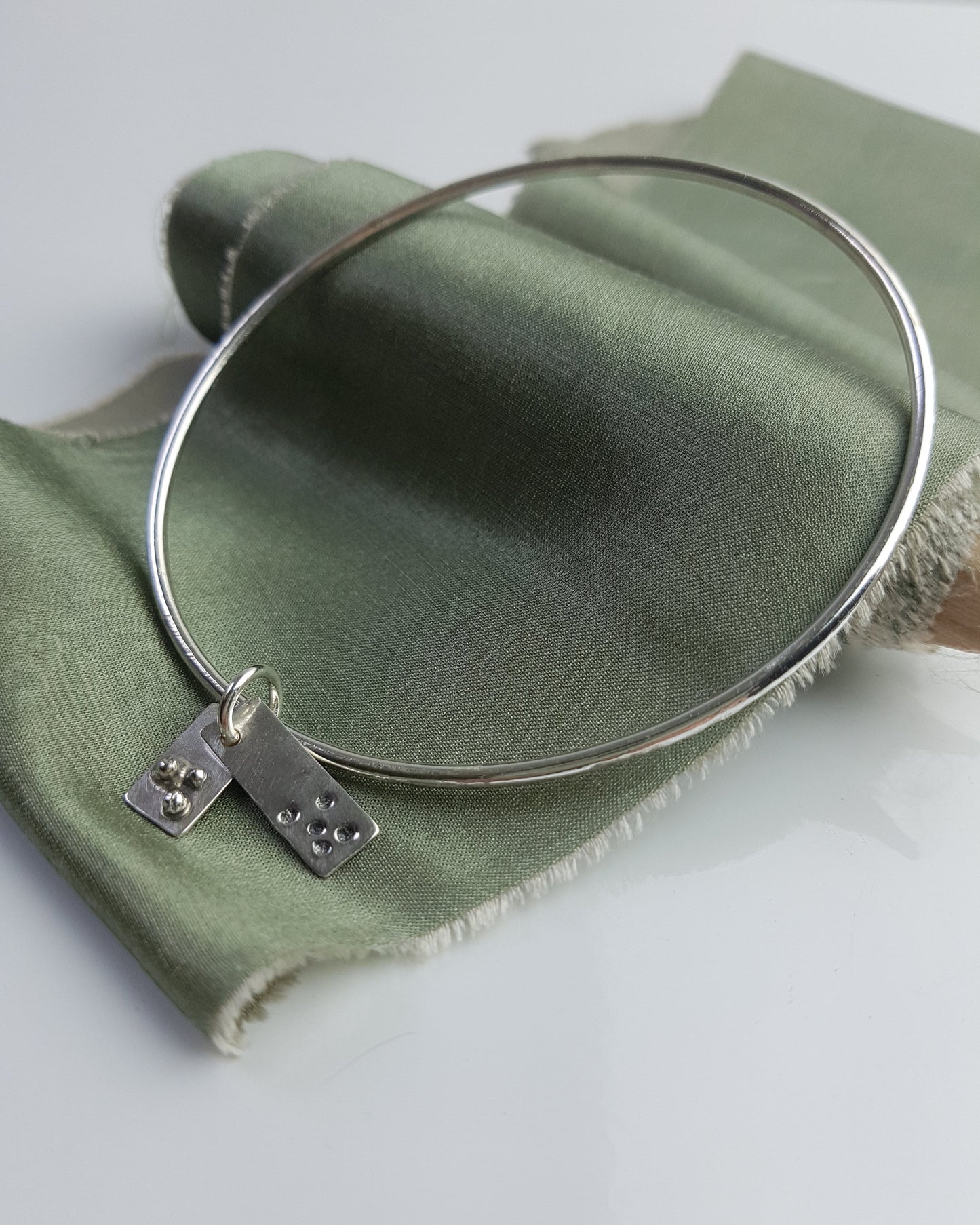 A silver bangle with two handmade rectangle charms rests on a green ribbon.