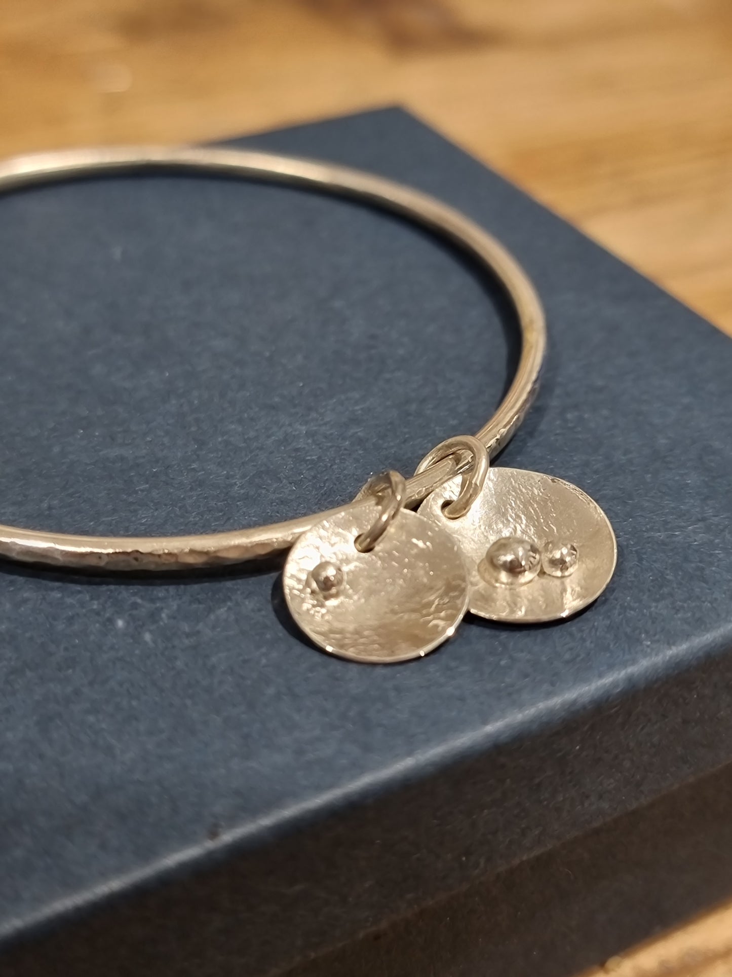 Silver bangle with two round disc charms on navy blue box.