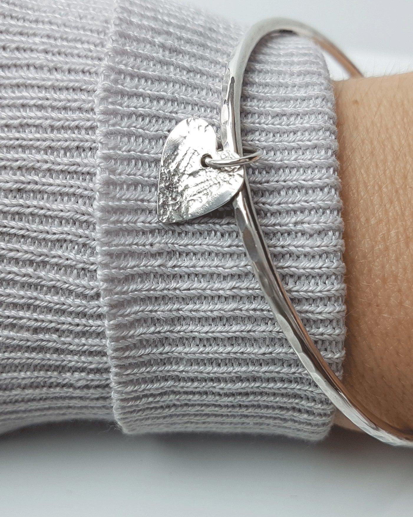 A woman's wrist with a grey cuff wearing a silver bangle with a heart shaped charm.